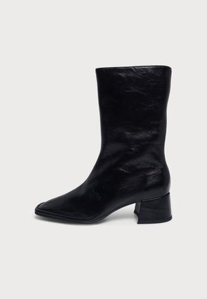 Botte en cuir noire mi-mollet à bout pointu et talon bloc bas sur un fond uni clair.