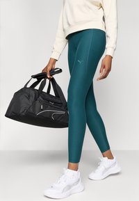 Leggings verde-água com uma textura suave, combinados com um sweatshirt creme. Mala de ginásio preta com múltiplos bolsos com zíper. Ténis brancos com detalhes em prata.