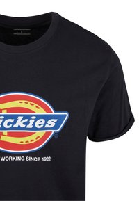 Juoda medvilninė marškinėliai su trumpomis rankovėmis, su spalvingu apvaliu logotipo dizainu ir tekstu „Dickies dirba nuo 1922“ priekyje.