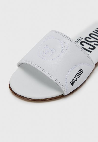 Sandale slide blanche en matériau synthétique avec broderie d'un ours en peluche et marquage noir. Texture lisse avec semelle plate.