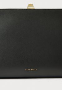 Mustast nahast käekott, millel on sile tekstuur, kuldse tooniga kinnitus ja minimalistlik disain. Brändinimi 'Coccinelle' on kuldse kirjaga reljeefne.