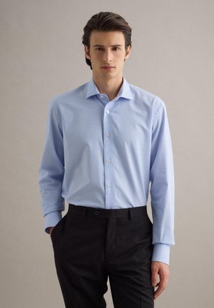 Junger Mann, der ein hellblau kariertes Hemd in schwarze Hosen steckt, steht mit einer Hand in der Tasche vor neutralem Hintergrund.