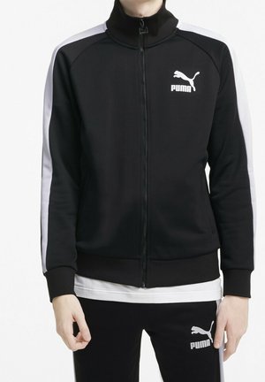 Chaqueta de chándal negra con cremallera y cuello alto, franjas laterales blancas y logotipo de Puma en el pecho. Tejido suave y diseño ajustado.
