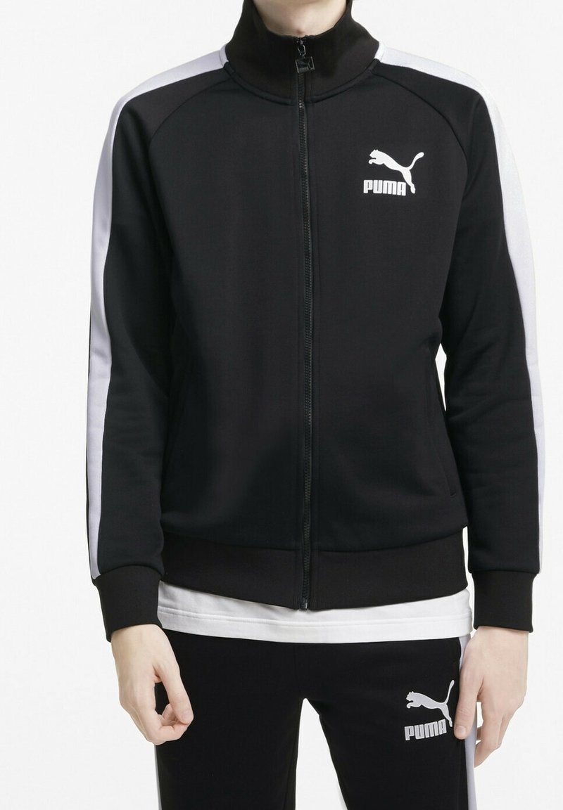 Giacca da allenamento nera con zip e colletto alto, strisce bianche sui lati e logo Puma sul petto. Tessuto liscio e design aderente.