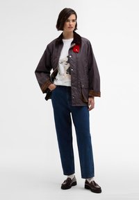 Casaco impermeável cinza com colarinho de veludo cotelê castanho, fecho com botões de pressão e forro xadrez, combinado com jeans azuis largos e mocassins castanhos.