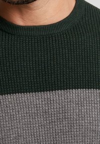 Gerippter Strickpullover mit einem grünen oberen Teil und einem kontrastierenden grauen unteren Teil, rundem Halsausschnitt und strukturiertem Stoff.