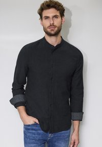 Camicia abbottonata in tessuto nero con colletto alla coreana, maniche lunghe e riflesso grigio sui polsini arrotolati. Texture liscia, vestibilità classica.
