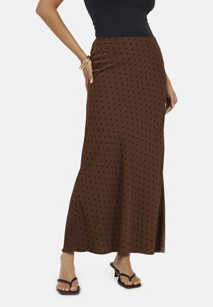 Vrouw draagt een lange bruine rok met zwarte stippen, een zwart mouwloos topje, zwarte sandalen met hakken en gouden armbanden aan de linkerpols.