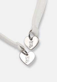 Δύο ασημένια μενταγιόν σε σχήμα καρδιάς με χαραγμένο το γράμμα "J" που είναι προσαρτημένα σε λευκές υφασμάτινες θηλιές.