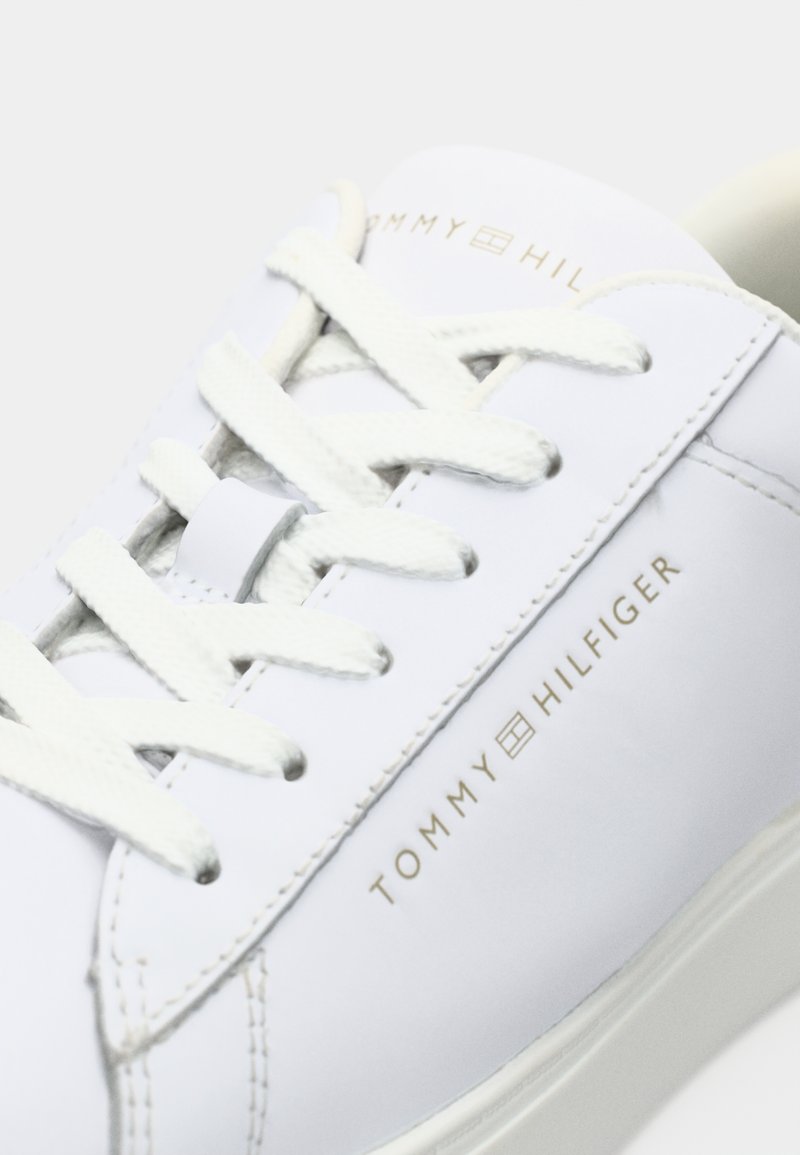 Weißer Sportschuh mit Obermaterial aus strukturiertem Leder, mit flachen weißen Schnürsenkeln und einem goldfarbenen, eingeprägten Logo an der Seite. Glatte Gummisohle.