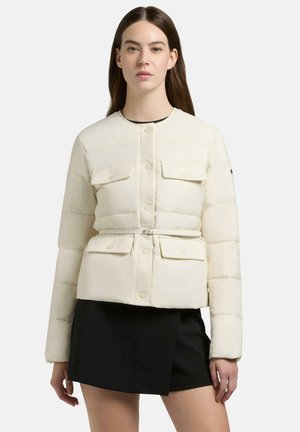 Vrouw met lang bruin haar, gekleed in een crèmekleurige pufferjas met riem en een zwarte asymmetrische minirok, tegen een witte achtergrond