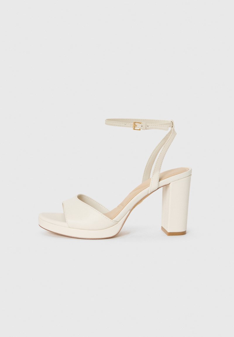 Sandalia blanca de tacón alto con tacón grueso, punta abierta, correa al tobillo y hebilla dorada, exhibida sobre un fondo claro y liso.