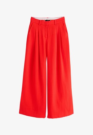 Weißes ärmelloses Top, in hoch taillierte, weite rote Hose mit Gürtelschlaufen und plissiertem Vorderteil gesteckt.