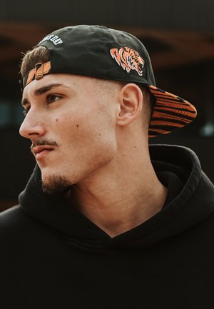 Cappello nero con grafica di una tigre e dettagli arancioni, indossato al contrario, abbinato a una felpa nera. Il profilo del soggetto è messo in risalto.