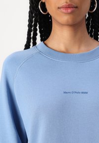 Gaiši zila sporta kapuce ar ribotu apaļu kaklu un raglanu piedurknēm; uz priekšas diskreta reljefa logo "Marc O’Polo DENIM".
