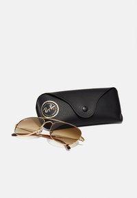 Ray-Ban aviator sunglasses with brown gradient lenses, gold metal frame, and temple tips in brown, accompanied by a black textured case.
