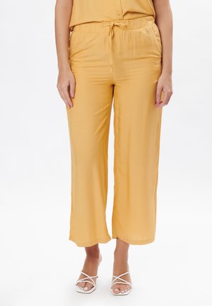 Gele, wijdvallende broek met een zachte textuur, elastische tailleband en verstelbare strik. Deze zijn gecombineerd met witte, gestrikte sandalen.