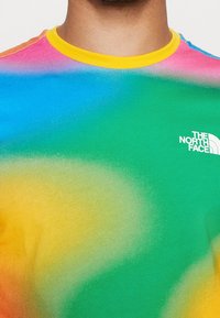 Detailní záběr na trup v multibarevném triku s batikováním, žlutým límečkem a bílým logem The North Face na zelené části.