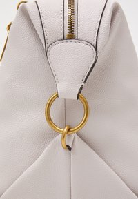 Sekcja torby z kremowej teksturowanej skóry z złotymi metalowymi pierścieniami i widocznym przeszyciem zamka.