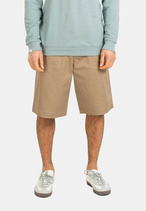 Hombre con pantalones cortos beige hasta la rodilla, sudadera azul claro y zapatillas blancas con gris, posando contra un fondo liso.