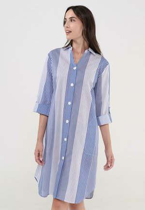 Mujer con vestido camisa de botones a rayas verticales azules y blancas hasta la rodilla y mangas tres cuartos, mirando hacia su izquierda.