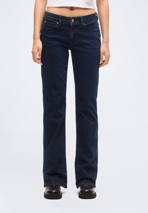 Személy sötétkék bootcut farmerben, fekete cipőben és egy rövid fehér felsőben áll, egy egyszerű világos háttér előtt.