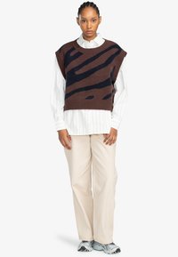 Brauner strickfreier Pullover mit dunkelblauen abstrakten Streifen, über einem weiß gestreiften Hemd getragen, kombiniert mit beigen Hosen und grauen Sneakers.