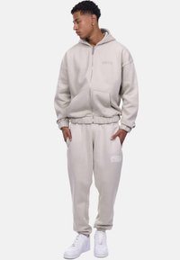 Light beige zip-up hoodie with kangaroo pockets and matching joggers. Both have a soft texture and feature printed accents. White sneakers.