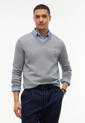Jeune homme portant un pull gris col en V par-dessus une chemise bleue boutonnée, un pantalon bleu marine à fines rayures et une ceinture noire, debout, une main dans la poche.