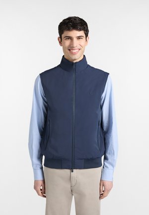 Mladý muž sa usmieva, nosí námornícky modrú vestu so zipsom cez svetlomodrú pruhovanú košeľu a béžové nohavice, stojí pred jednoduchým bielym pozadím.