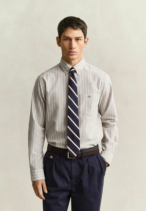 Jeune homme portant une chemise beige rayée à boutons, une cravate rayée bleu marine et jaune, et un pantalon bleu foncé, debout devant un fond uni.