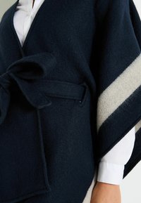 Poncho de lana en un oscuro color navy con una amplia franja crema, que presenta un cierre delantero con lazo y un acabado suave y texturizado. Se ven puños de camisa blanca debajo.