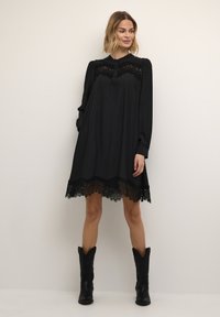 Schwarzes Langarmkleid mit Spitzeneinsatz, ausgestellt geschnitten und mit einer filigranen Spitzenkante. Kombiniert mit schwarzen Knöchelstiefeln.