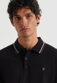Schwarzes Poloshirt mit einer Knopfleiste mit drei Knöpfen, kontrastierendem gestreiften Kragen und einem goldenen Sticklogo auf der linken Seite.