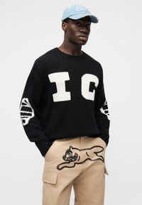 Μαύρο πλεκτό πουλόβερ με άσπρα γράμματα "IC" και ριγωτά στοιχεία στα μανίκια, σε συνδυασμό με μπεζ παντελόνια τύπου cargo με σχέδιο μαύρης γάτας.