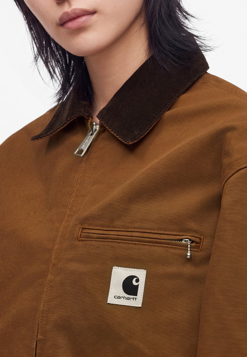 Young person wearing brown work jacket with dark brown corduroy collar, silver zipper, and white Carhartt logo patch on chest pocket.