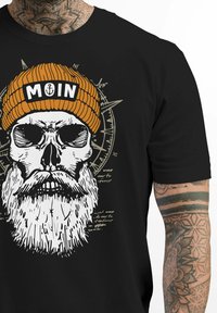 Schwarzes T-Shirt mit einem Grafikdesign eines Schädels, der eine orange gestreifte Mütze trägt. Der Text lautet "MOIN". Das Design enthält einen Kompass und handgeschriebene Notizen.