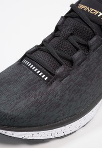 Chaussure de sport noire avec tige en mesh texturé, semelle intermédiaire blanche tachetée contrastante et lacets noirs avec des accents réfléchissants subtils.
