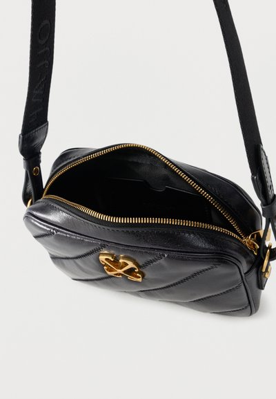 Μαύρη καπιτονέ δερμάτινη τσάντα χιαστί με χρυστό φερμουάρ και διακοσμητικό χρυσό έμβλημα, μερικώς ανοιχτή με ρυθμιζόμενο υφασμάτινο λουρί.