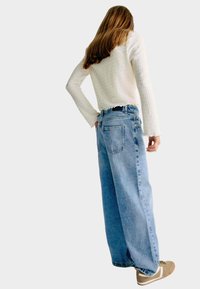 Jeans anchos de lavado claro con cintura alta, combinados con una blusa blanca texturizada de manga larga y zapatillas beige.