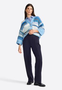 Cardigan lavorato a maglia blu con strisce chiare e scure, taglio oversize, scollatura a V e chiusura con bottoni, abbinato a pantaloni blu scuro e scarpe nere.