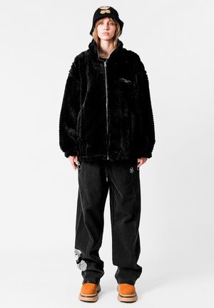 Chaqueta de piel sintética negra con cuello alto y cremallera, combinada con pantalones negros holgados con detalles únicos y zapatos color canela.