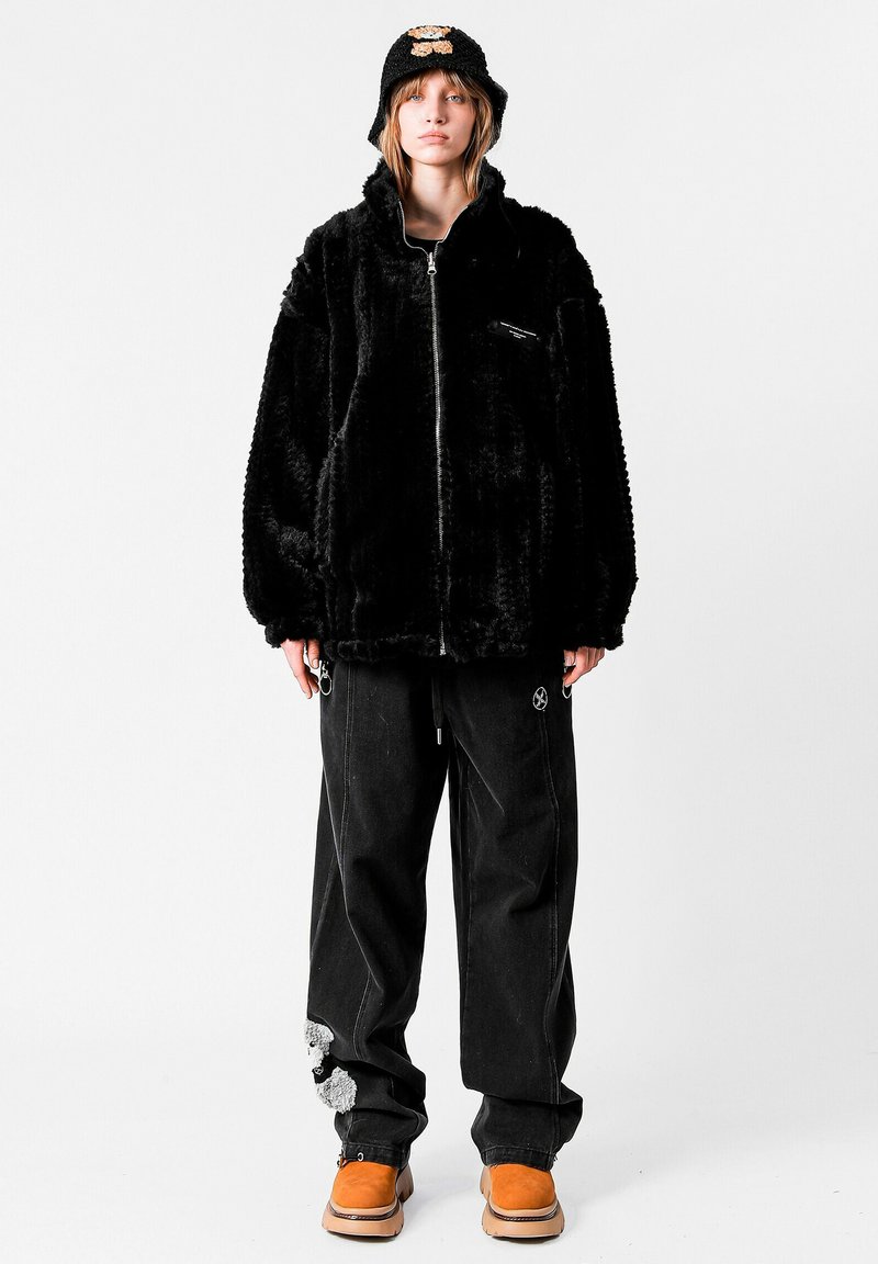 Chaqueta de piel sintética negra con cuello alto y cremallera, combinada con pantalones negros holgados con detalles únicos y zapatos color canela.