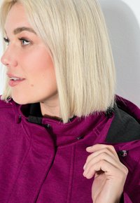 Blondynka w magentowej kurtce poprawia kołnierz, patrząc w lewo na tle jednolitego białego tła.