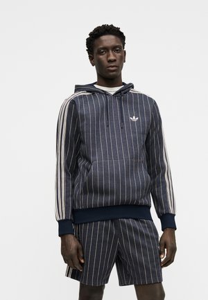 Mand iført en mørkeblå hoodie med striber og matchende shorts, stående foran en ensfarvet lys grå baggrund.