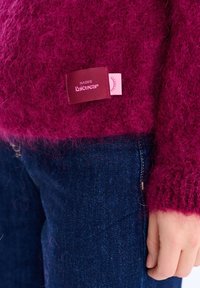 Zbliżenie na dłoń spoczywającą blisko brzegu puszystego, magentowego swetra dzianego z metką "NAOKO Knitwear" na ciemnoniebieskich dżinsach.