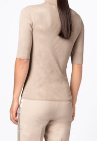 Beżowy prążkowany top z wysoką wykładanym kołnierzem i krótkimi rękawami, prezentujący dopasowaną sylwetkę oraz fakturowany materiał. Tylny widok podkreśla gładki krój.