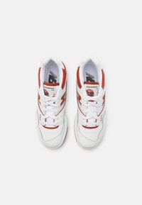 Paire de baskets New Balance 550 blanches et rouges avec lacets noués, vue de dessus sur un fond clair uni.