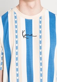 Randig t-shirt i blått, krämfärgat och vitt. Har vertikala ränder med ett lövmönster. Svart broderad text "Kara" på bröstet.