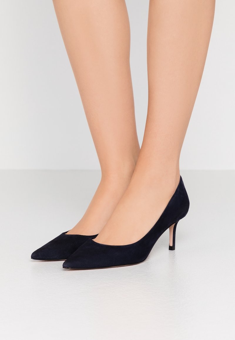 Jambes portant des escarpins en daim noir à bout pointu et talon kitten sur un sol blanc et un mur gris clair en arrière-plan.